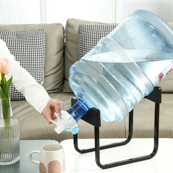 Person pouring water from a large plastic container into a cup on a table with a couch in the background.